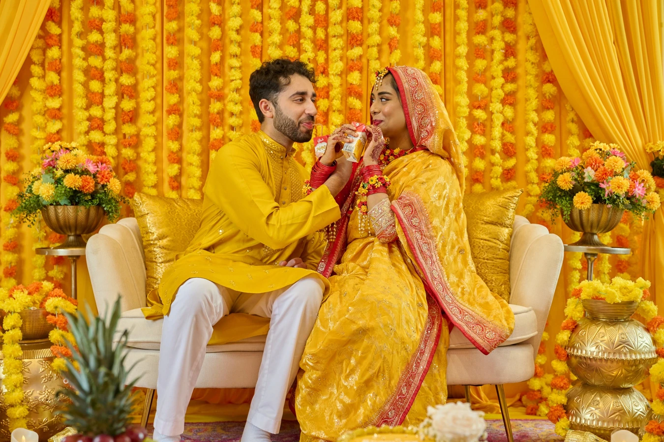 Bride and groom in yellow Haldi attire sharing a Maaza juice box in front of a marigold flower curtain